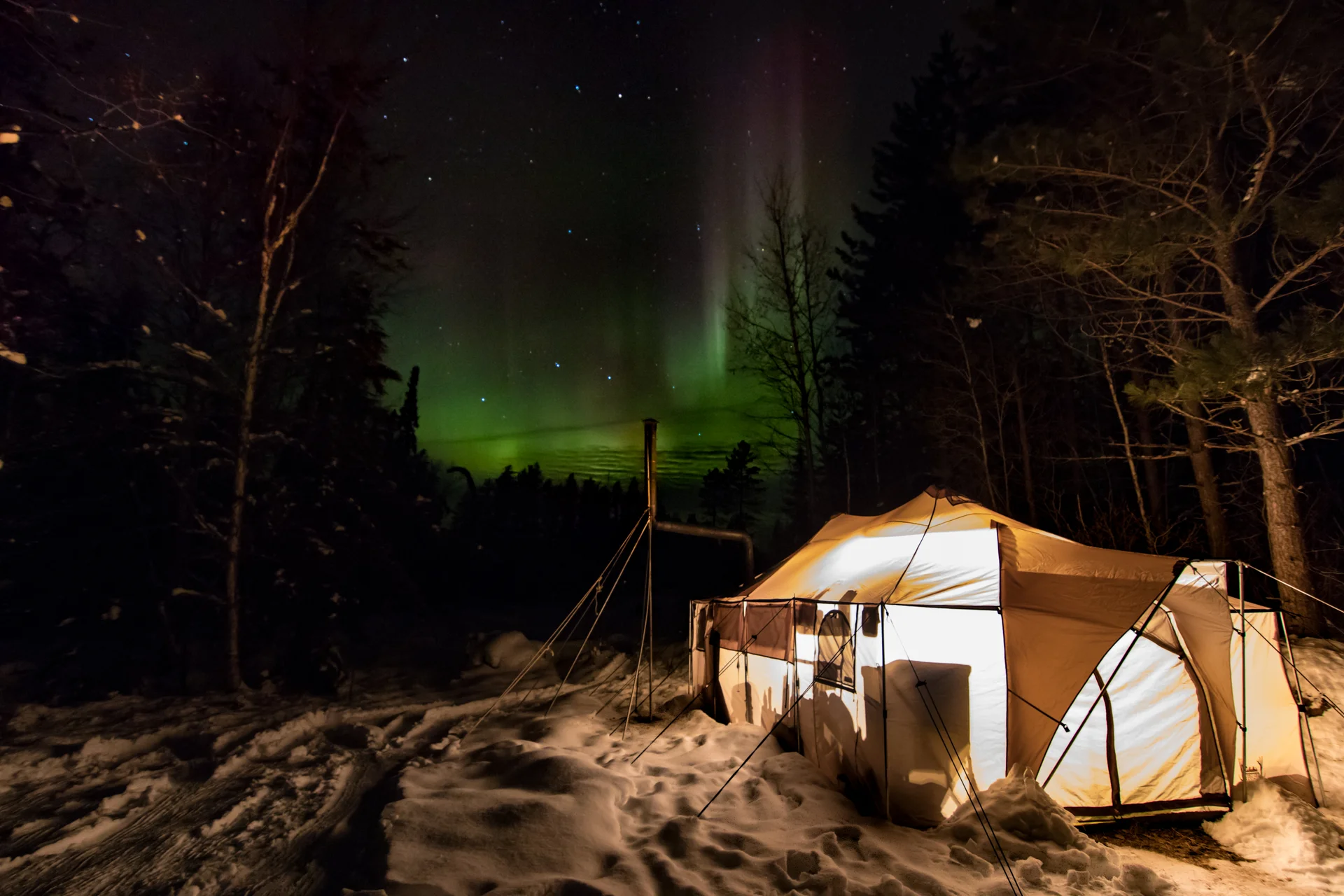 Northern lights aurora borealis glowing green over a lit canvas tent in winter forest