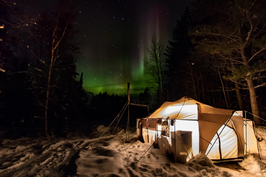 Northern lights aurora borealis over lit canvas tent in winter forest