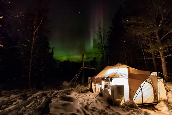 Aurora borealis glowing green over a lit canvas tent in winter forest