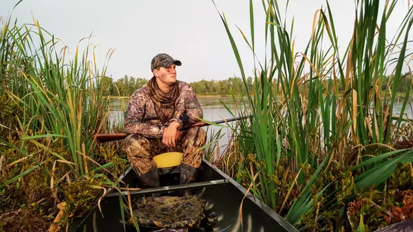 Duck hunter in camo gear sitting in canoe among marsh reeds